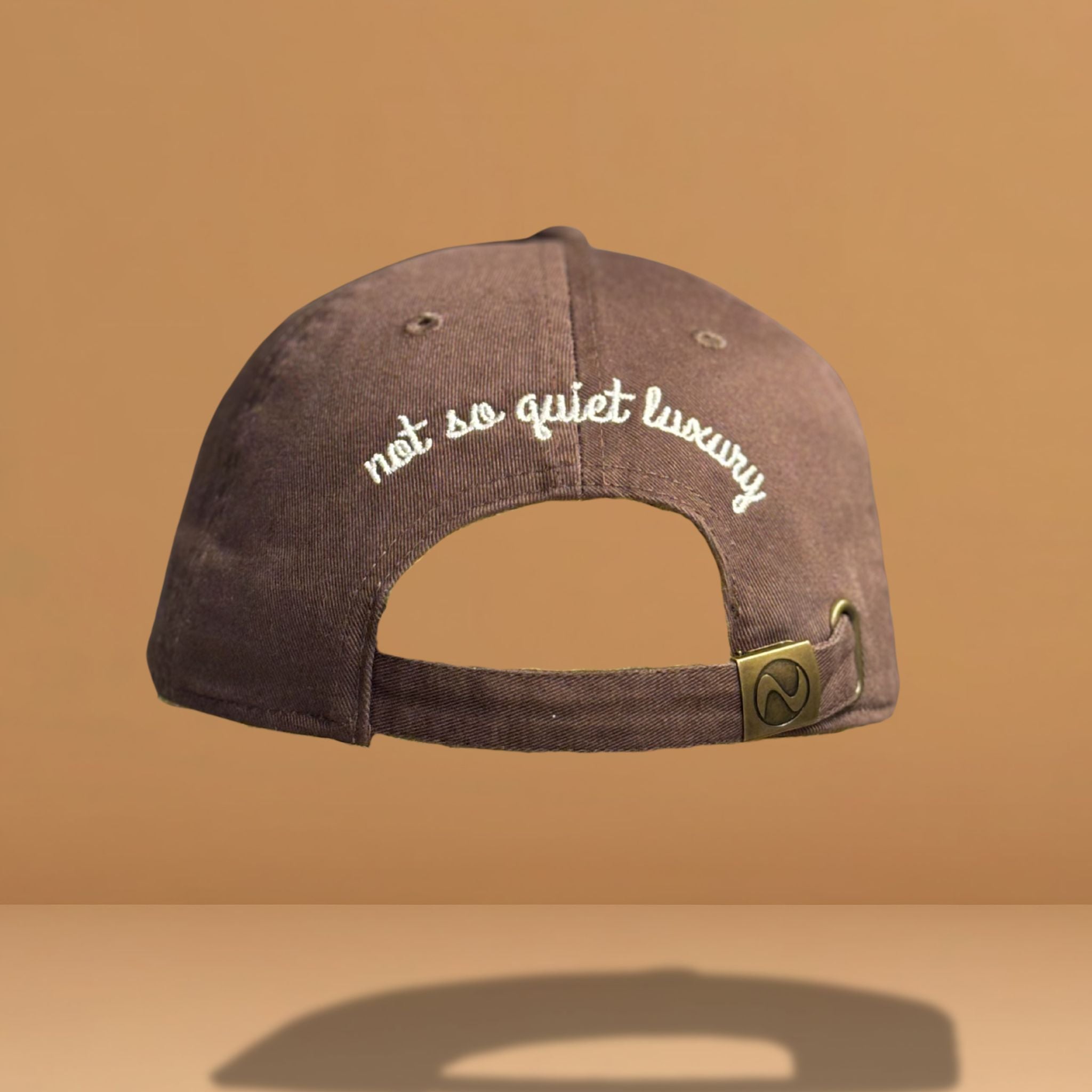 Back view of brown cotton baseball cap showing white embroidered text 'not so quiet luxury' across the back panel with adjustable strap closure and metal hardware, photographed against warm tan background.