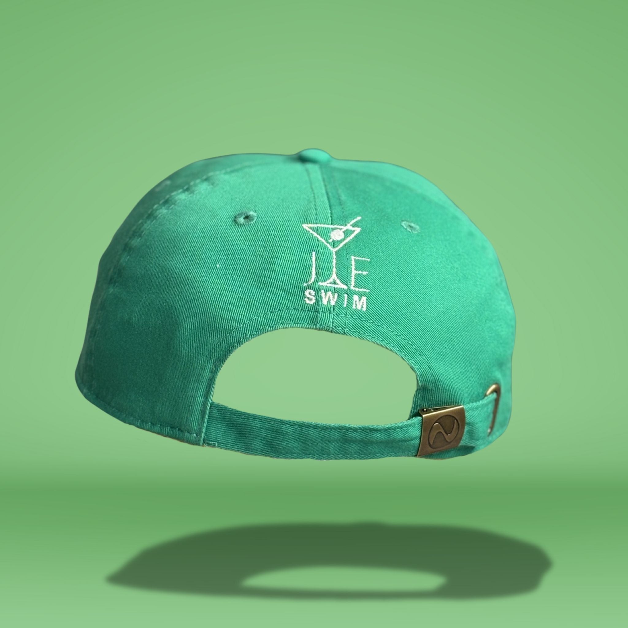 Back view of green cotton baseball cap showing white embroidered 'JE SWIM' logo with martini glass icon and adjustable strap closure with metal hardware, photographed against bright green background.
