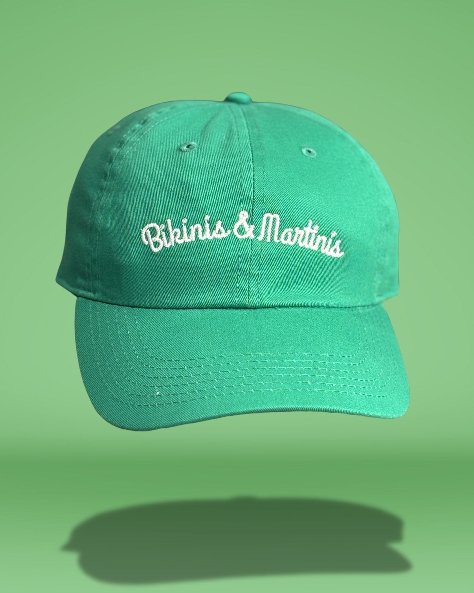 Green cotton baseball cap with white embroidered 'Bikinis & Martinis' script text across the front, featuring curved brim and classic six-panel construction, photographed against bright green background.