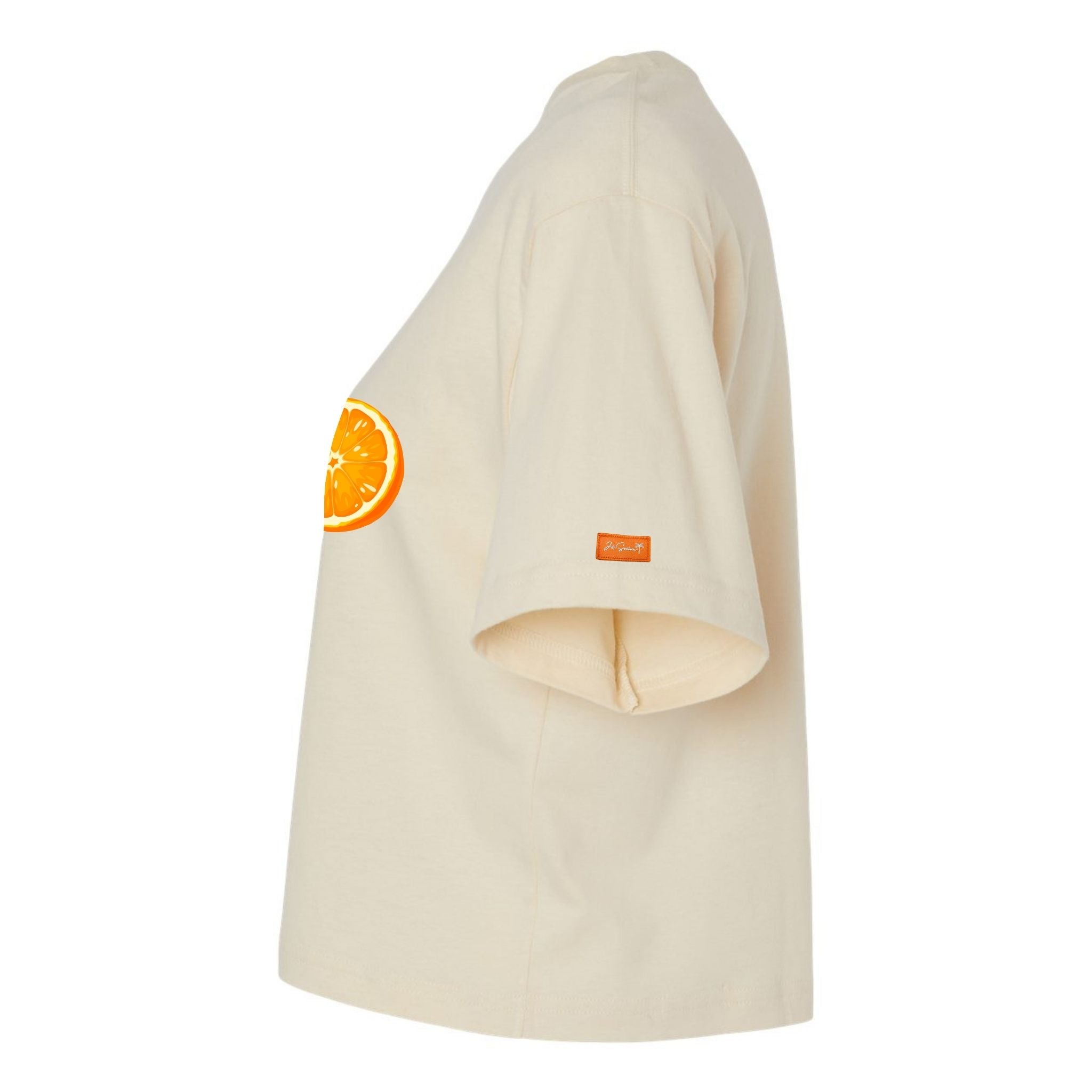 Side view of Je swim cream colored cropped t-shirt with orange slice graphic on the chest and orange 'JE' logo label on the sleeve, displayed on a clean white background.