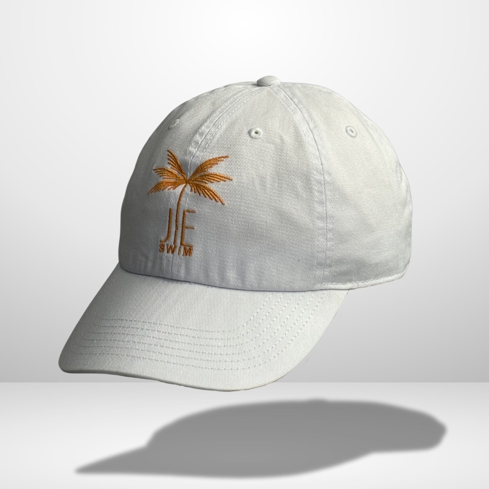 Side angle view of white cotton baseball cap with orange embroidered palm tree and 'JE SWIM' logo, featuring curved brim and classic six-panel construction, photographed against clean white background.