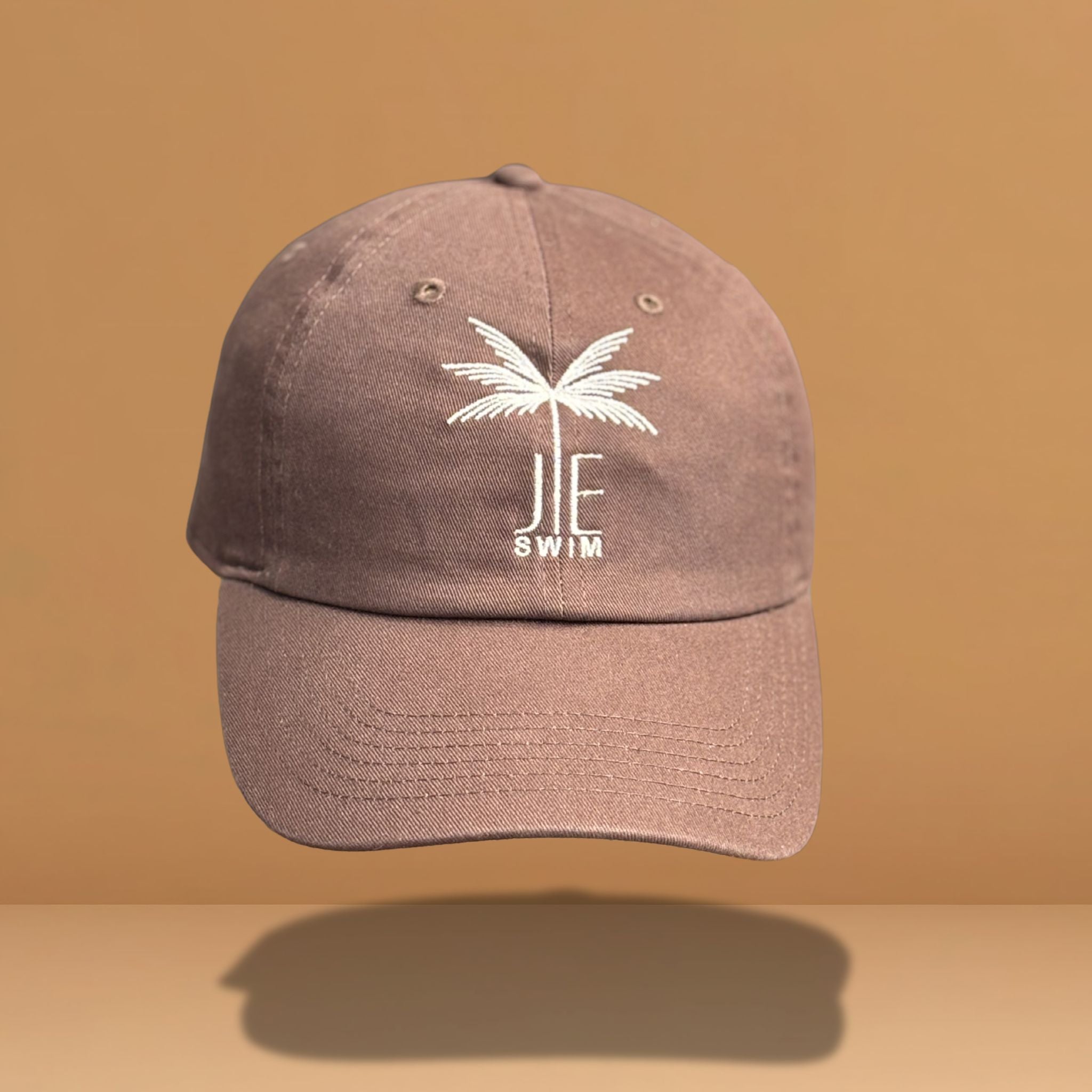 Brown cotton baseball cap with white embroidered palm tree and 'JE SWIM' logo on front, featuring curved brim and classic six-panel construction, photographed against warm tan background.