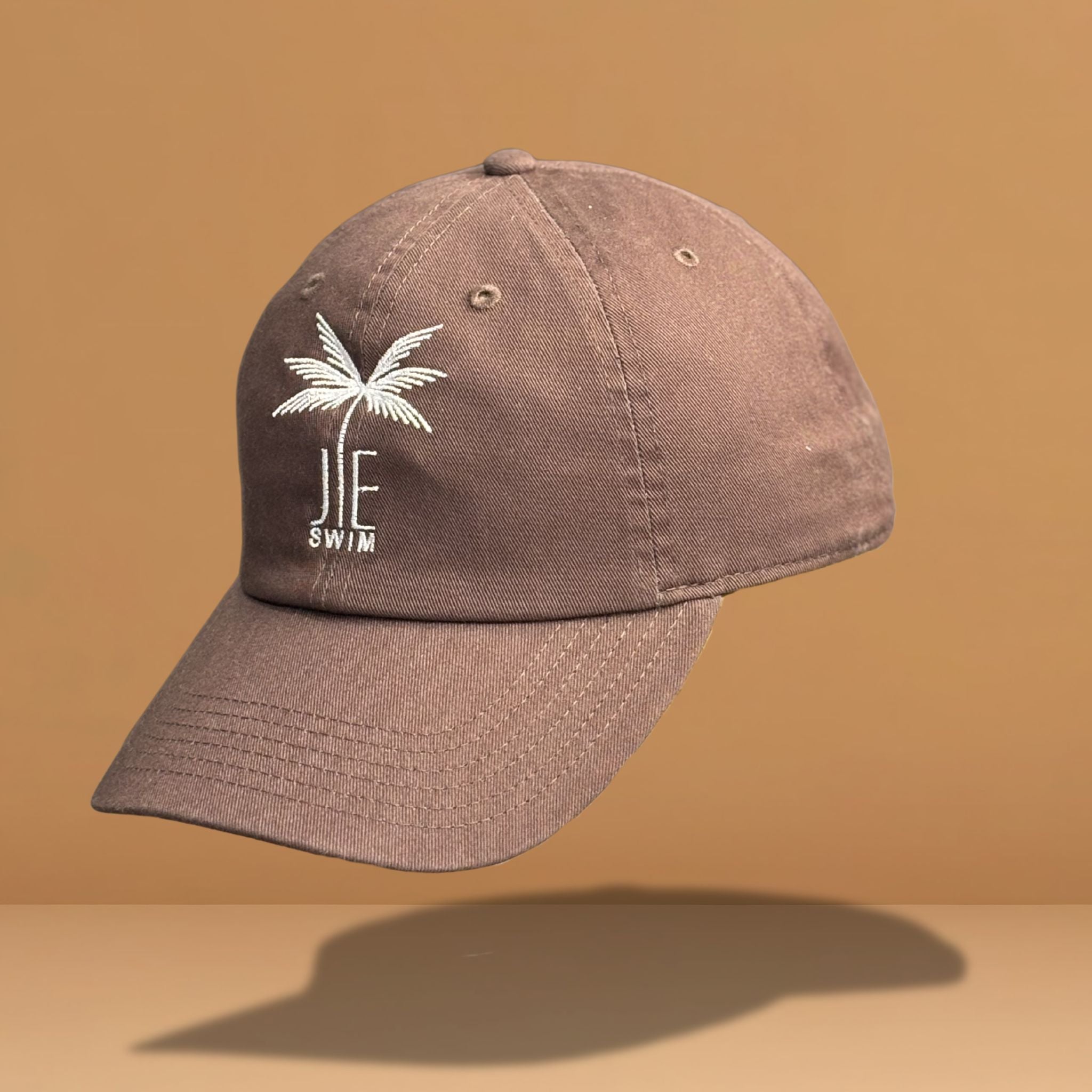 Side angle view of brown cotton baseball cap displaying white embroidered palm tree and 'JE SWIM' logo, showing the hat's profile and curved brim structure, photographed against warm tan background.