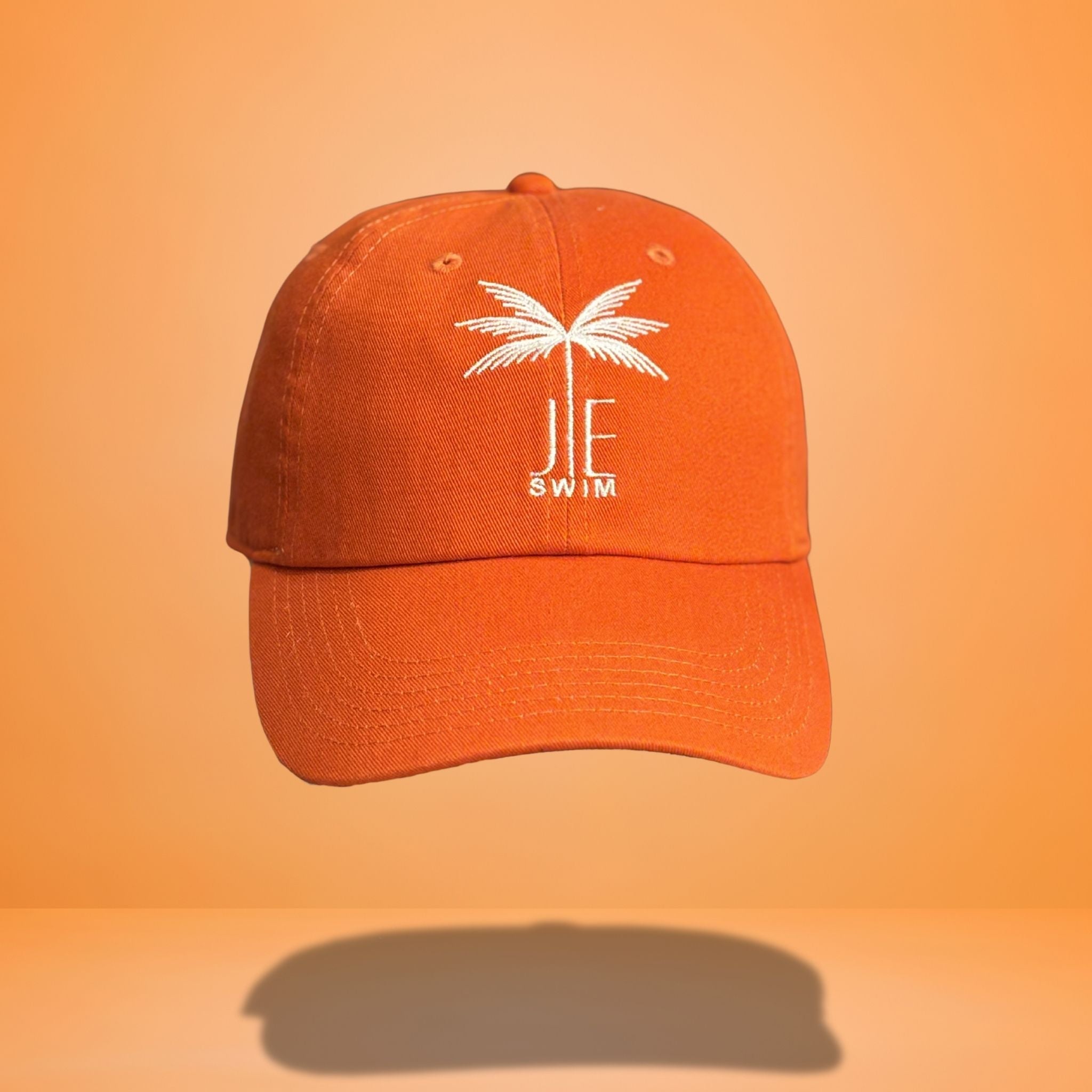Orange cotton baseball cap with white embroidered palm tree and 'JE SWIM' logo on front, featuring curved brim and classic six-panel construction, photographed against bright orange background.
