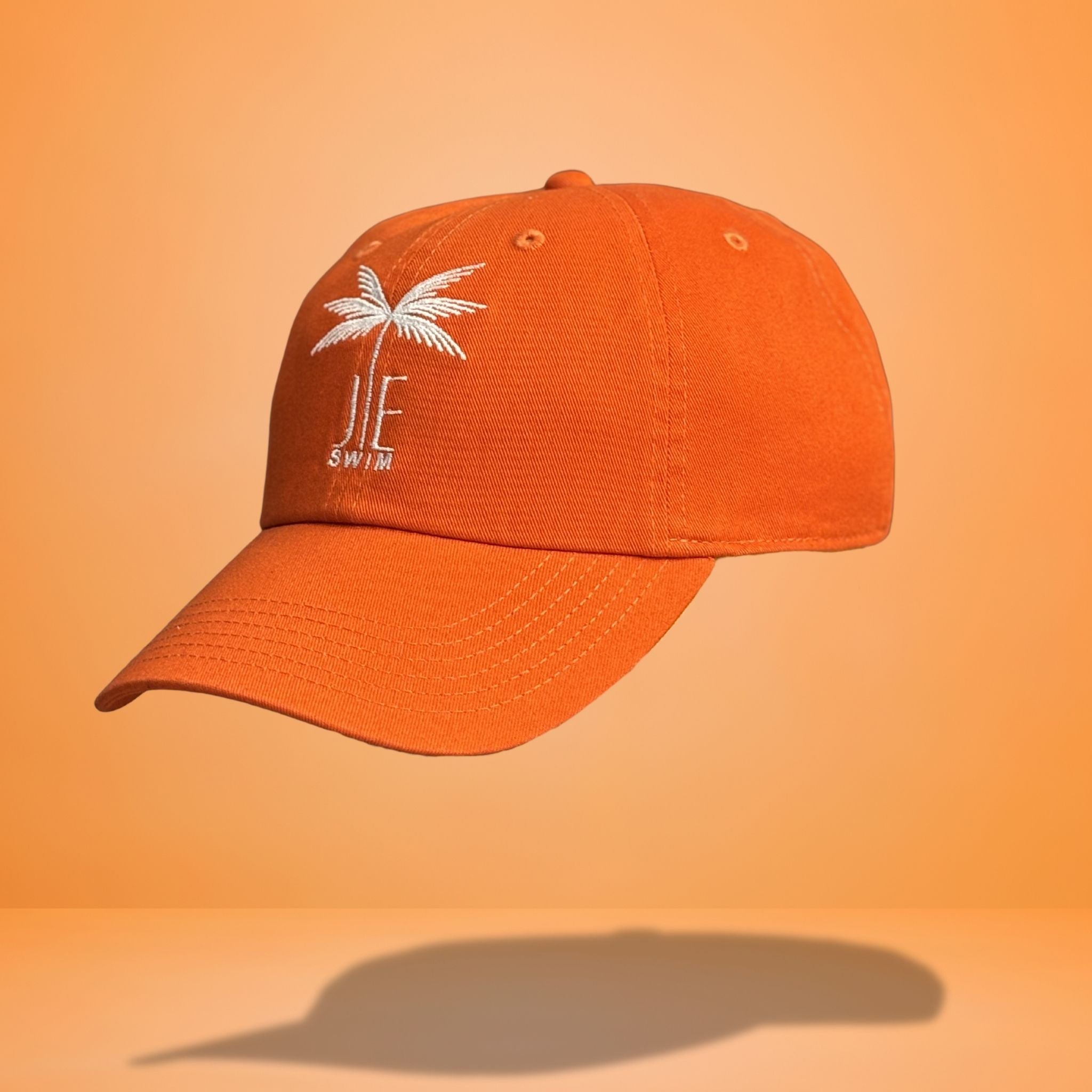 Side angle view of orange cotton baseball cap showing white embroidered palm tree and 'JE SWIM' logo with curved brim profile, photographed against bright orange background.