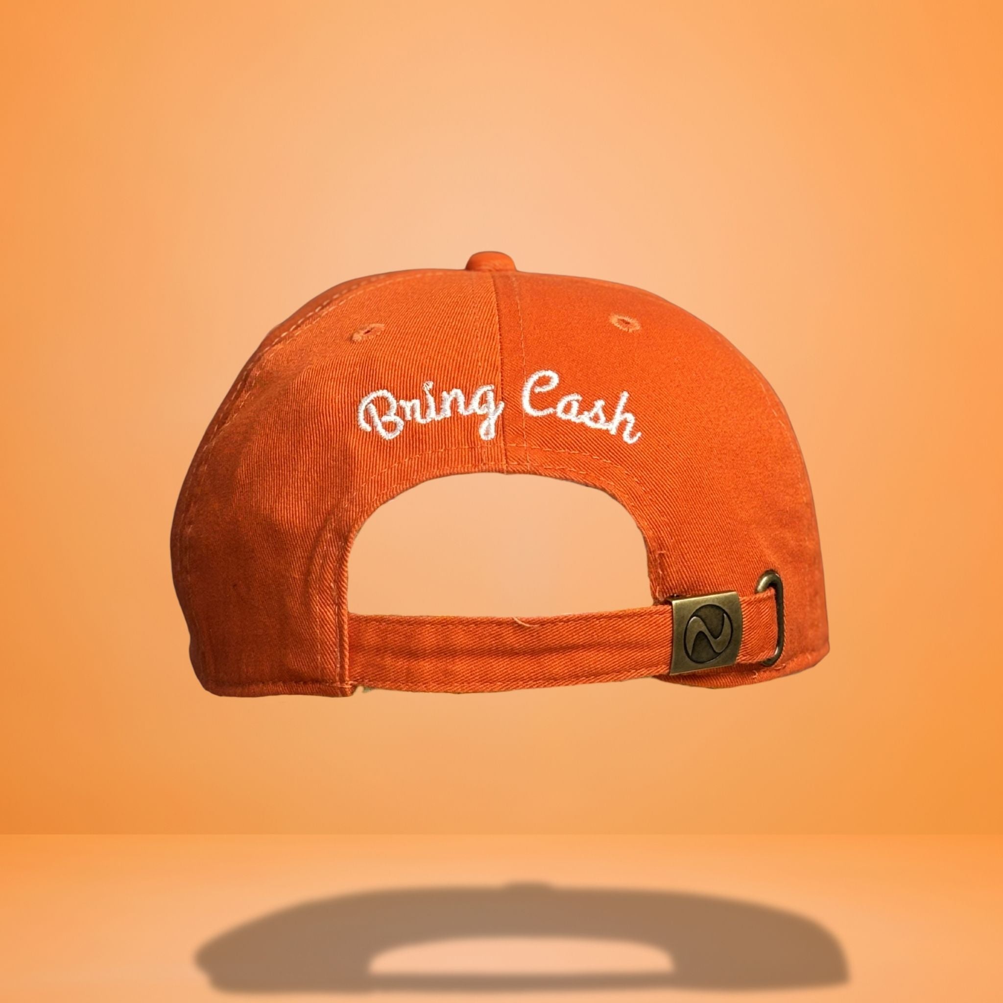 Back view of orange cotton baseball cap showing white embroidered 'Bring Cash' script text across the back panel with adjustable strap closure and metal hardware, photographed against bright orange background.