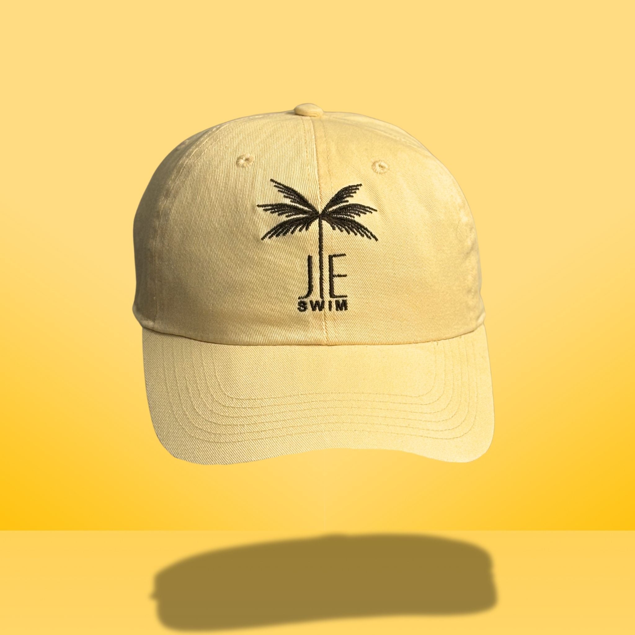 Yellow cotton baseball cap with black embroidered palm tree and 'JE SWIM' logo on front, featuring curved brim and classic six-panel construction, photographed against bright yellow background.