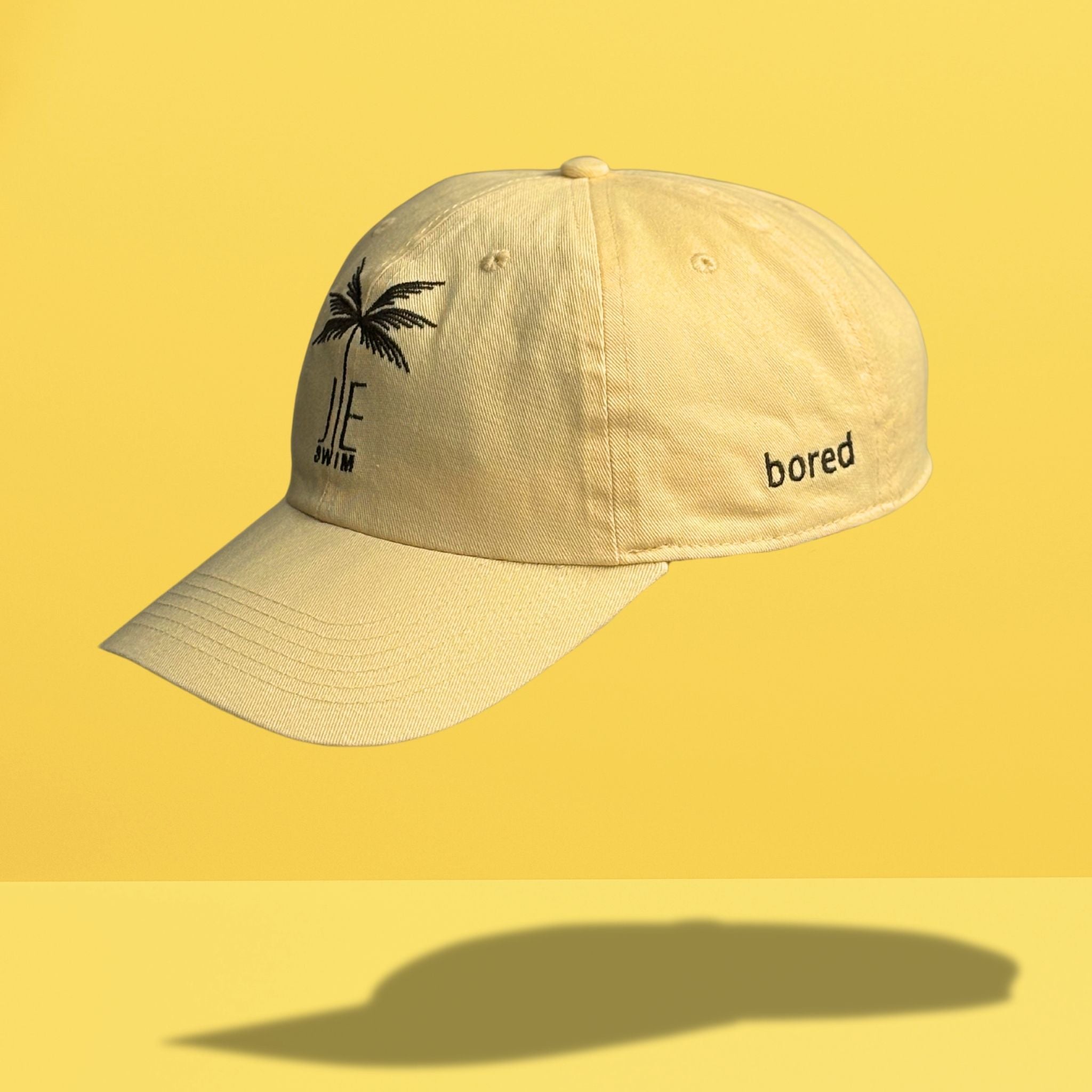 Side angle view of yellow cotton baseball cap showing black embroidered palm tree and 'JE SWIM' logo on front with 'bored' text embroidered on the side panel, photographed against bright yellow background.