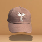 Brown cotton baseball cap with white embroidered palm tree and 'JE SWIM' logo on front, featuring curved brim and classic six-panel construction, photographed against warm tan background.