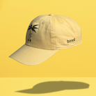 Yellow JE Swim baseball cap with "Bored" embroidered on the side and a black palm tree logo.